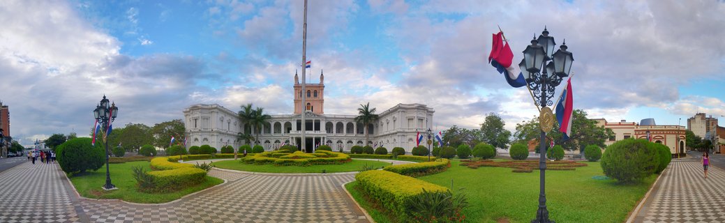 White neoclassical presidential palace with central tower, flanked by flag-adorned lampposts and manicured gardens with checkered walkways under cloudy sky