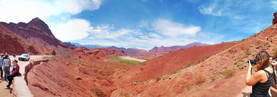 Wide panoramic landscape showing red rock canyon formations with tourists at viewpoints, white SUV parked nearby, valley with sparse vegetation, distant mountains under blue sky with white clouds in Cafayate, Argentina