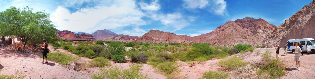 Panoramic view of red rock mountain formations in Cafayate desert with green shrubs, blue sky with clouds, and tourists near a white van