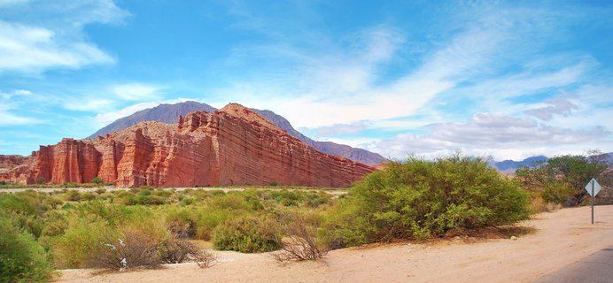 Red layered rock cliffs with vertical striations in a desert landscape with green shrubs and blue sky in Cafayate, Argentina