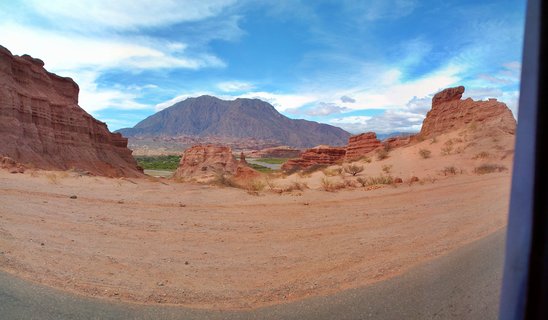 Desert landscape showing red sandstone rock formations and cliffs with a large mountain in the background under a blue sky with white clouds in Cafayate, Argentina