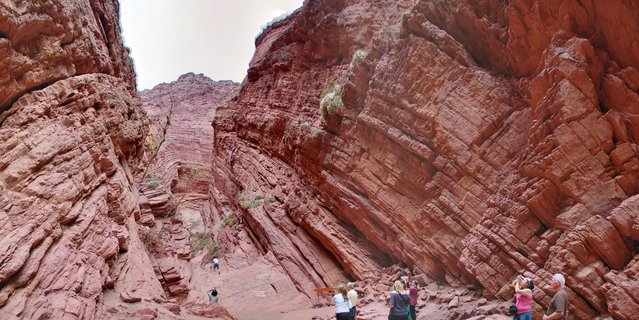 Low-angle view of tourists standing among towering red sandstone rock formations with diagonal layered patterns in a narrow canyon
