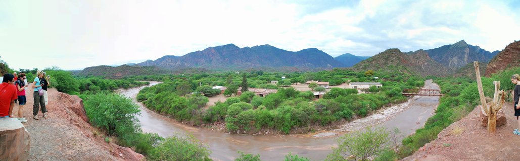 Panoramic view of a winding river valley with green trees, mountains in the background, and tourists standing on viewing platforms on both sides