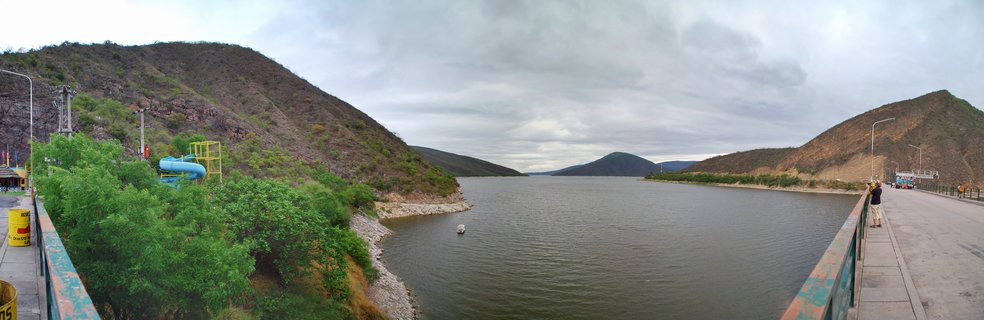 Panoramic view of Dique Los Sosa reservoir near Cafayate, Argentina, showing calm blue water between brown arid hills under overcast sky, with dam structure, water slide facility, and walkway with people visible