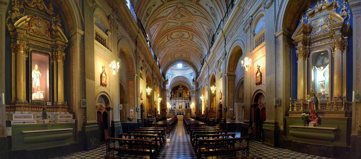 Interior view of a colonial cathedral showing ornate golden altarpieces on both sides, vaulted painted ceiling, checkered floor, wooden pews, and religious statues in Salta, Argentina