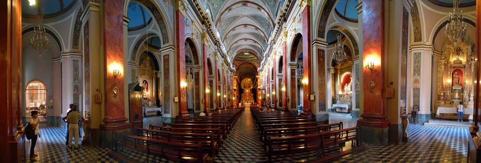 Panoramic interior view of an ornate cathedral with red marble columns, golden altars, crystal chandeliers, vaulted frescoed ceilings, wooden pews, and checkered floor with visitors walking inside