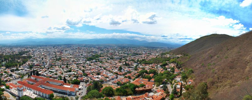 Panoramic cityscape of Salta, Argentina showing red-tiled roof buildings stretching across a valley with mountains in the background and a hillside on the right under a blue sky with white clouds