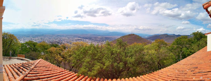 Panoramic landscape showing a city valley view from above with orange terracotta roof tiles in foreground, green trees in midground, urban sprawl in the distance, and mountains under a blue sky with white clouds