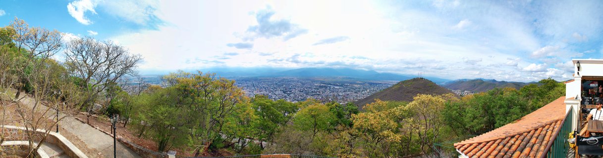Panoramic landscape showing Salta city in a valley surrounded by mountains, with trees and colonial architecture in the foreground under a blue sky with clouds