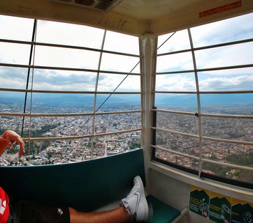 Interior view from cable car showing person's legs in white sneakers, metal safety bars, and panoramic cityscape of Salta with mountains beyond