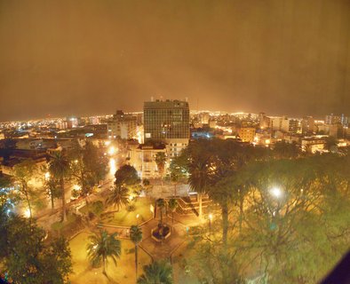 Night aerial photograph of Salta, Argentina showing an illuminated city plaza with palm trees in the foreground, a prominent glass high-rise building in the center, and urban lights extending to the horizon under an overcast sky