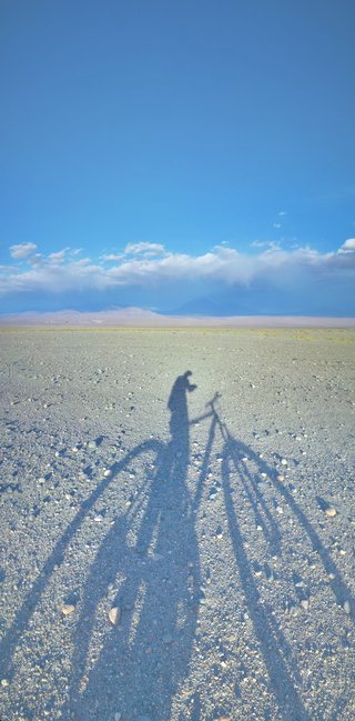Shadow of a photographer with tripod cast on rocky desert ground, with mountains and clouds in the distance under blue sky