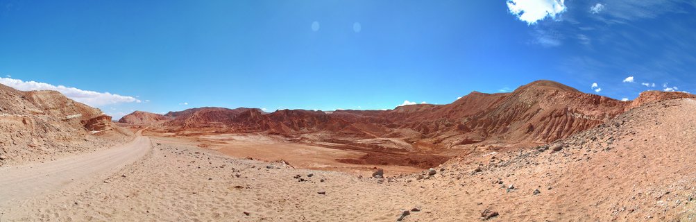 Wide desert landscape showing red rock formations and eroded hills with a dirt road curving through sandy terrain under a bright blue sky with scattered white clouds in the Atacama Desert