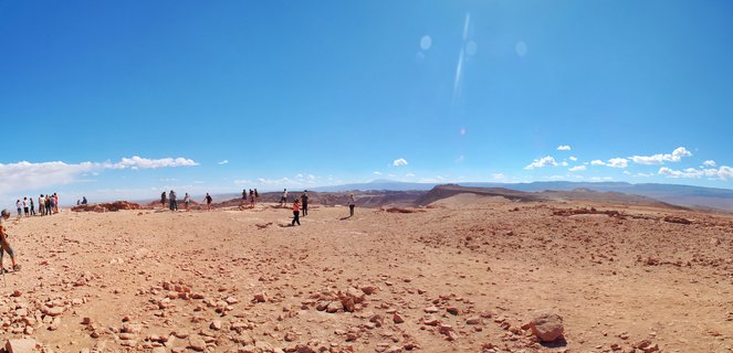 Wide desert landscape showing reddish-brown rocky terrain with approximately 20 tourists walking and standing, clear blue sky with scattered white clouds, distant mountains on horizon