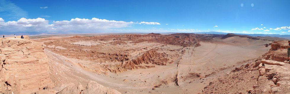 Wide panoramic photograph of a desert canyon landscape with red-orange rock formations, eroded valleys, and distant mountains under a bright blue sky with white clouds; small human figures visible on cliff edges