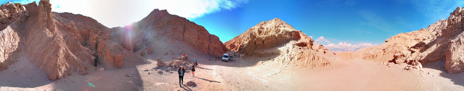 Panoramic view of reddish-brown eroded rock formations in a desert landscape with tourists walking on sandy terrain under a clear blue sky in Valle de la Luna, Chile