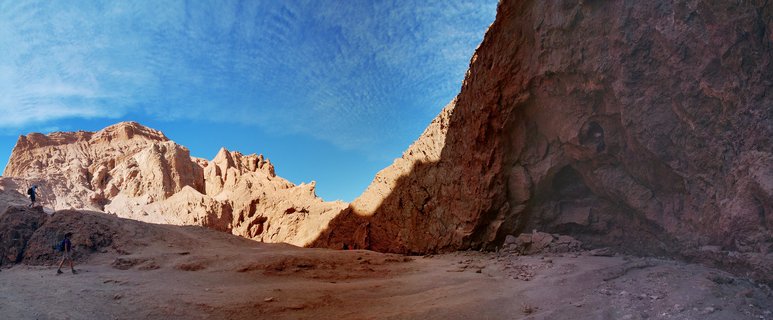 Two hikers walking through a desert canyon with tall reddish-brown rock formations and blue sky with clouds in San Pedro de Atacama, Chile