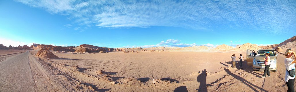 Panoramic view of tourists standing beside a silver van on a dirt road in the Atacama Desert near San Pedro de Atacama, Chile. Reddish-brown eroded rock formations and sandy terrain extend across the landscape under a blue sky with wispy clouds.