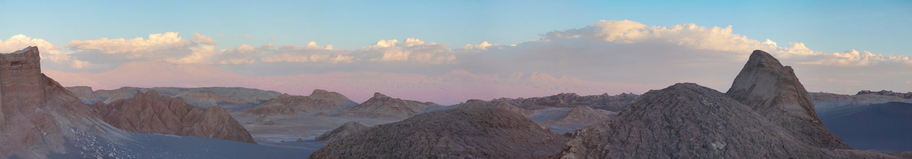Desert mountain landscape showing rocky formations and distant peaks under blue sky with scattered clouds at sunset