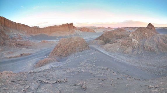 Panoramic desert landscape showing eroded salt and clay rock formations with layered cliffs in the background under a blue to pink gradient sky at sunset in Chile's Atacama Desert