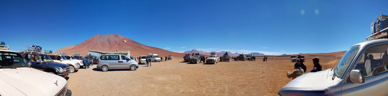 Panoramic view of multiple tour vehicles parked in a high-altitude desert with people standing nearby, a red volcanic mountain in the background, and clear blue sky above the barren landscape
