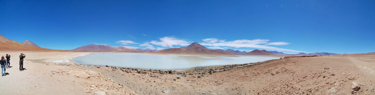 Panoramic view of turquoise lake surrounded by reddish-brown desert with three people standing on the shore, volcanic mountains in background under blue sky