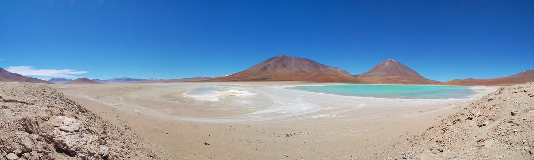 Wide panoramic landscape showing a turquoise salt lagoon surrounded by white mineral flats with two reddish-brown volcanic mountains under a clear blue sky in the Atacama Desert