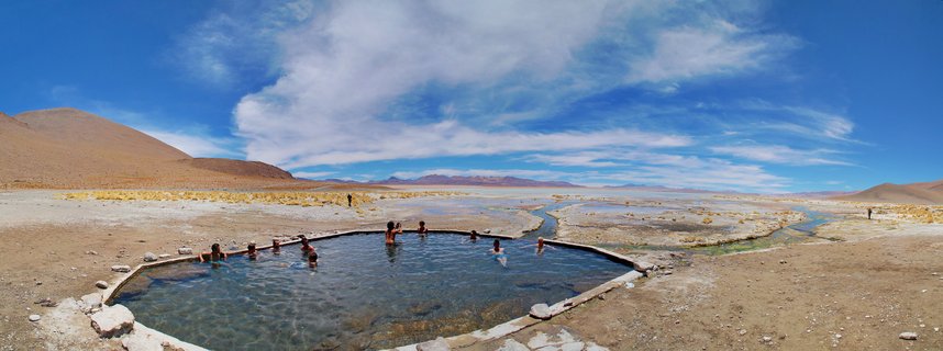 People bathing in a circular natural hot spring pool in a desert landscape with mountains, blue sky and white clouds in the background
