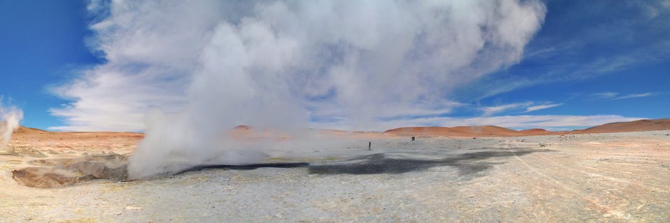 White steam billows from geothermal fumaroles across a pale mineral desert landscape with reddish-brown hills in the distance under a bright blue sky with scattered clouds.