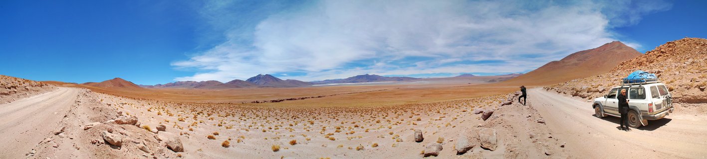 Panoramic desert landscape showing a white SUV with roof cargo parked on a dirt road in Bolivia's Altiplano region, with two people nearby, sparse vegetation, distant reddish mountains and volcanoes under blue sky with clouds