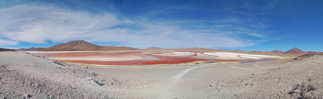 Panoramic view of Laguna Colorada showing red-colored salt lake water with white mineral deposits, surrounded by arid desert terrain and brown mountains under blue sky with scattered clouds