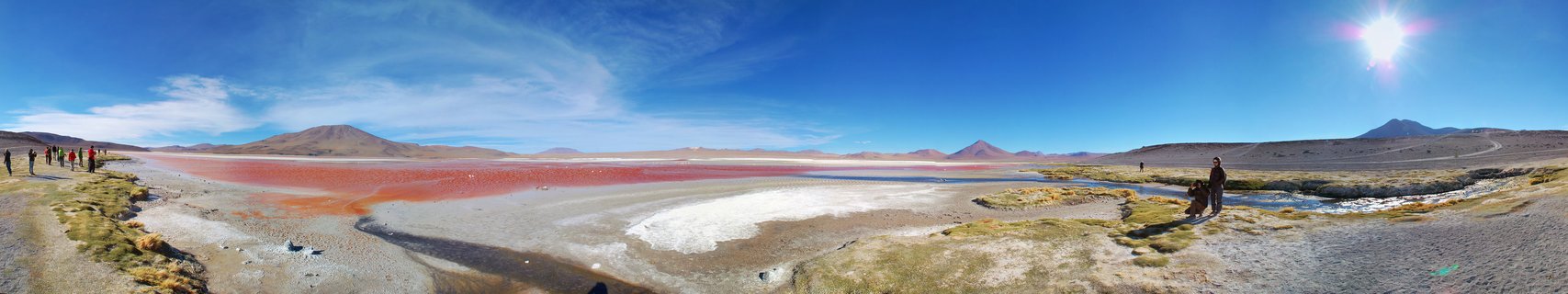 Panoramic view of Laguna Colorada showing red-colored salt lake water, white mineral deposits, arid terrain, distant mountains, blue sky with sun, and tourists walking along dirt paths on both sides of the frame