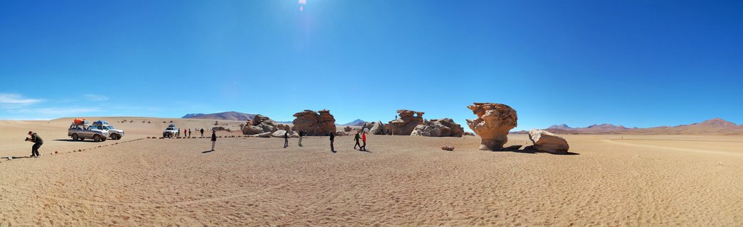 Panoramic view of wind-eroded rock formations in a desert landscape with tourists walking and two 4WD vehicles under a clear blue sky