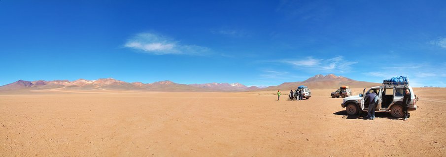 Three off-road vehicles with travelers stopped in a vast desert landscape with mountains in the background under a clear blue sky