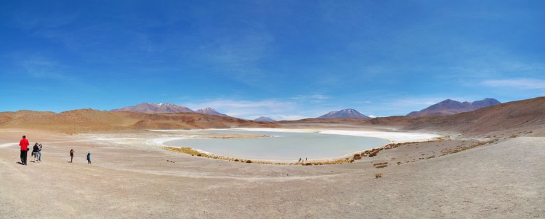 Wide desert landscape showing a turquoise salt lake surrounded by brown arid terrain with several tourists walking near the shore and volcanic mountains with snow caps in the distance under a clear blue sky