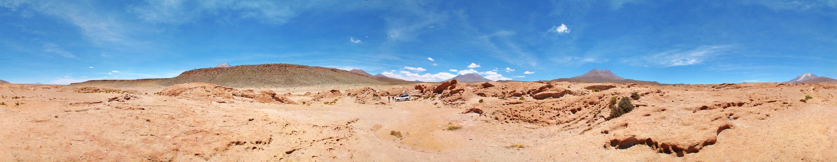 Wide desert landscape showing reddish-brown rocky terrain under blue sky with white clouds, distant mountains and volcanoes on horizon, small white vehicle visible in middle distance