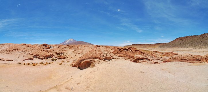 Desert landscape with red rock formations in foreground, sparse vegetation, clear blue sky with wispy clouds, and snow-capped mountain volcano in distance