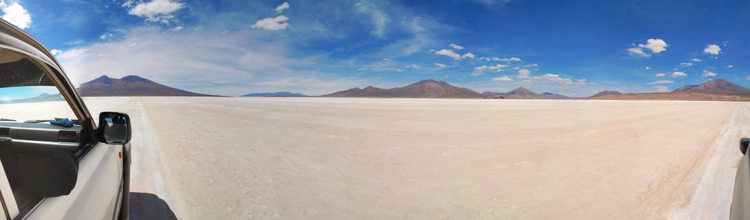 Panoramic view of white salt flats with partial view of white vehicle on left side, distant brown mountains on horizon, blue sky with white clouds above
