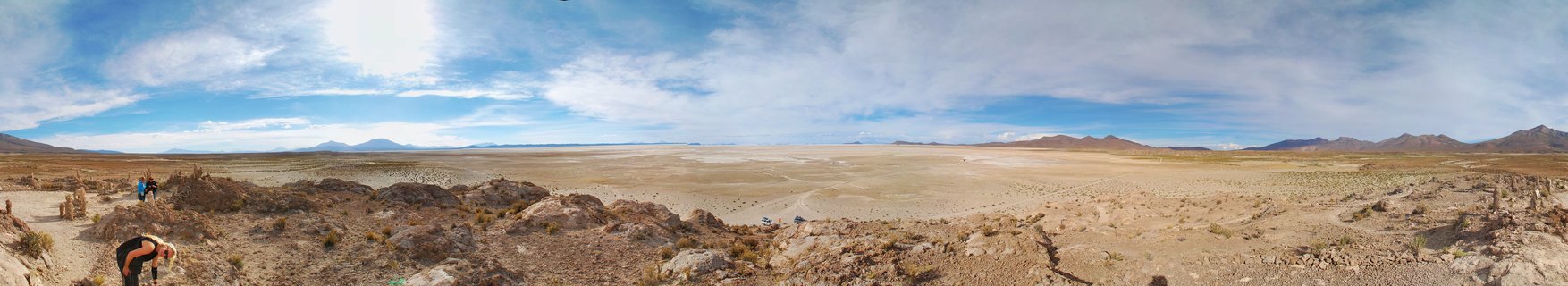 Wide panoramic desert landscape showing rocky foreground with two people, vast flat plain extending to distant mountains under blue sky with white clouds