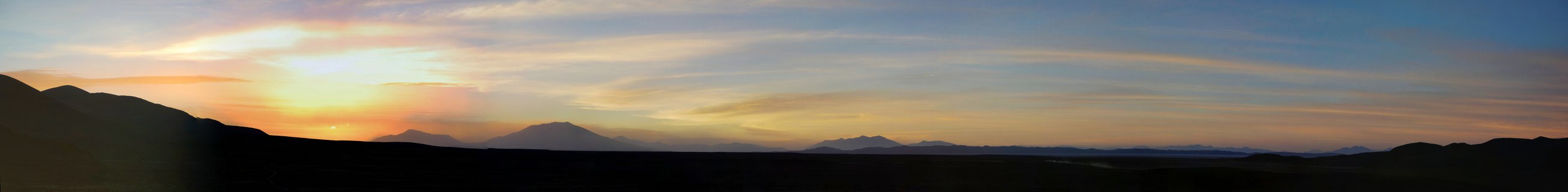 Panoramic view of dark mountain silhouettes against a colorful sunset sky with orange, yellow and blue horizontal cloud bands over the Atacama Desert in Bolivia.