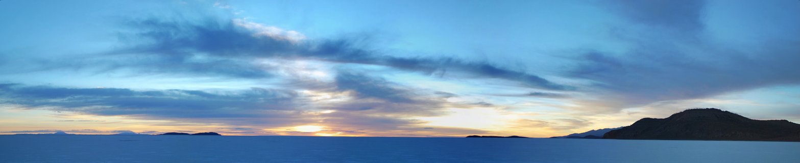 Panoramic landscape showing sunrise over vast salt flats with blue sky, scattered clouds, and dark mountain silhouettes on the horizon in Bolivia