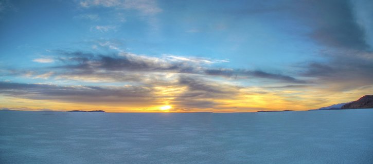Panoramic view of sunrise over a vast white salt flat with golden horizon, blue sky with clouds, and distant mountain silhouettes in Bolivia