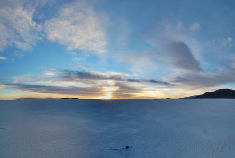 Aerial view of vast white salt flat at sunrise with golden sun on horizon, dramatic blue sky with clouds, and distant dark mountains in Bolivia