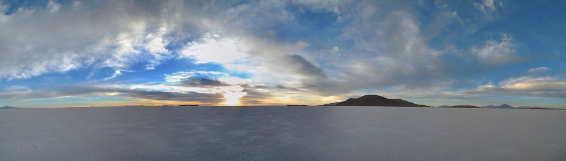 Panoramic view of sunrise over a vast white salt flat with mountains on the horizon and dramatic clouds filling a blue sky