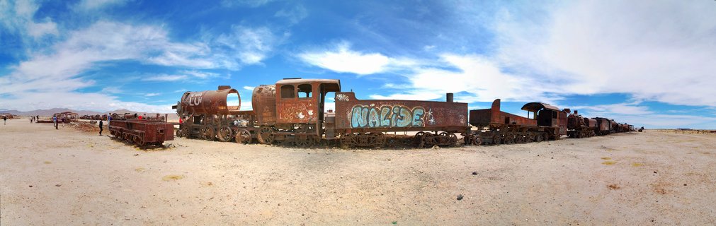 Rusted abandoned steam locomotives and train cars with graffiti sit in a desert landscape under blue sky with white clouds, tourists walking in the distance at Train Cemetery near Uyuni, Bolivia