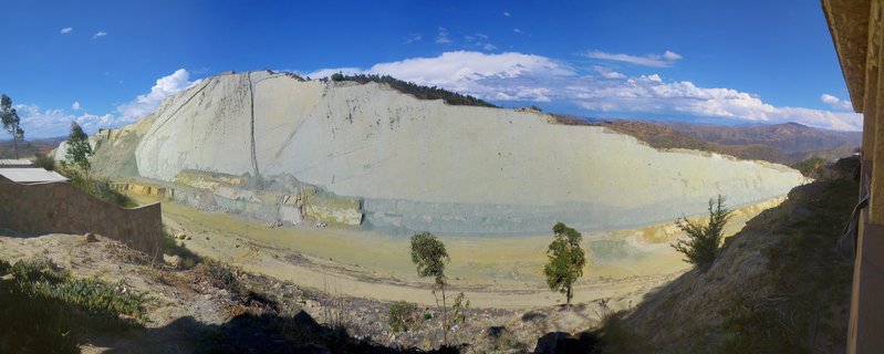 Wide view of a massive white limestone quarry cliff face with visible rock layers, sparse trees in foreground, blue sky with clouds above, mountain ranges in distance