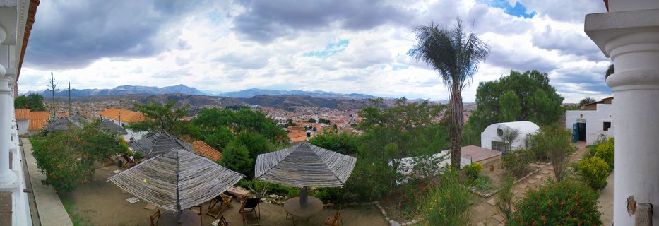 Panoramic view from a terrace with thatched umbrellas and white columns overlooking Sucre, Bolivia's colonial city with red-tiled roofs and distant mountains under cloudy sky