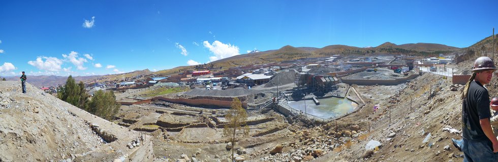 Wide panoramic photograph of a mining operation in Potosi, Bolivia showing industrial buildings, processing ponds, two workers wearing hard hats, arid brown hills, and blue sky with scattered white clouds