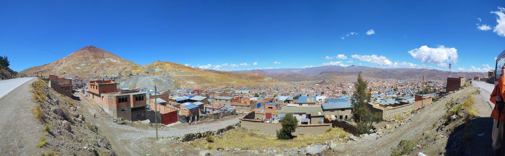 Wide panoramic photograph of Potosí, Bolivia showing densely packed city buildings with red and brown roofs beneath the prominent Cerro Rico mountain under clear blue sky