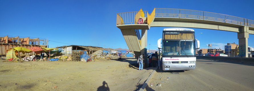 A white bus parked under a concrete pedestrian bridge with yellow railings, people boarding, colorful market stalls with tarps to the left, clear blue sky, dirt ground, Potosí Bolivia